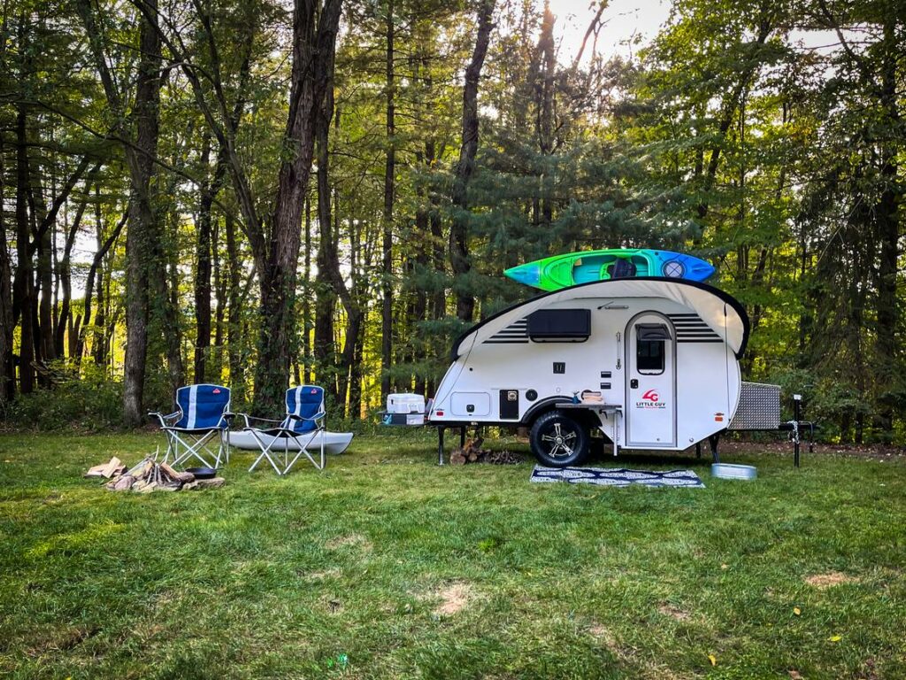 A Little Guy Trailers Micro Max set up at a grassy campsite with lawn chairs and a kayak on top.