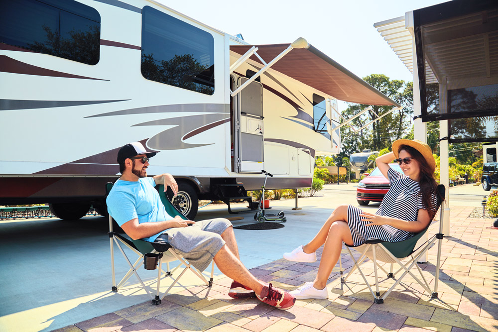 A couple sits outside of their fifth wheel during their summer RV travels