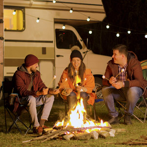 A group of friends roast marshmallows around a campfire outside an RV