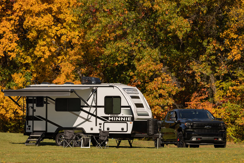 A truck is hitched to a 2023 Winnebago Micro Minnie in front of fall trees. It is one of the best RVs for 2023.